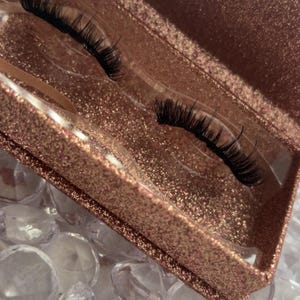 Puede incluir: Un par de pestañas postizas negras exhibidas en una caja de purpurina dorada rosada abierta. Las pestañas están sobre un inserto de plástico transparente. El fondo presenta cristales claros y facetados.