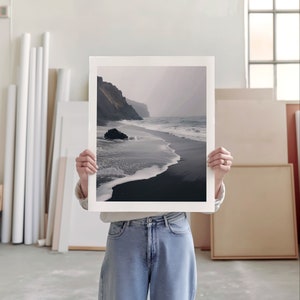 An artist studio room interior. A woman artist holds up a poster in front of her face. Concrete floor. Paper rolls and blank art canvas. Large warehouse windows, sunlight streams in