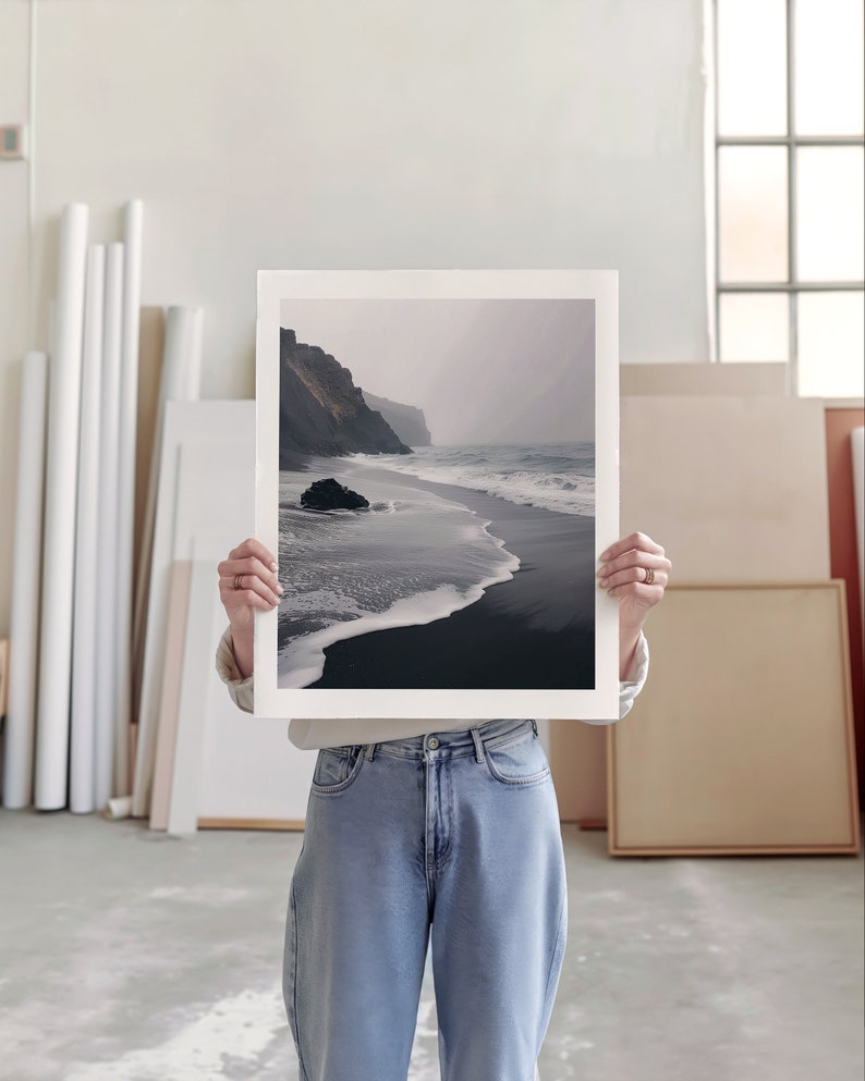 An artist studio room interior. A woman artist holds up a poster in front of her face. Concrete floor. Paper rolls and blank art canvas. Large warehouse windows, sunlight streams in