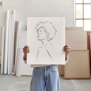 An artist studio room interior. A woman artist holds up a poster in front of her face. Concrete floor. Paper rolls and blank art canvas. Large warehouse windows, sunlight streams in