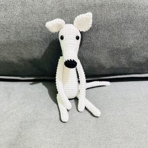 May include: A white crochet dog with a black nose and eyes. The dog has large, floppy ears and is sitting with its legs outstretched. The toy is made of soft yarn. A charming, handmade item.