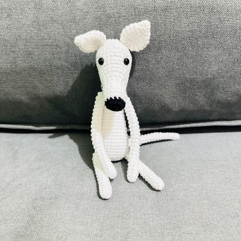 May include: A white crochet dog with a black nose and eyes. The dog has large, floppy ears and is sitting with its legs outstretched. The toy is made of soft yarn. A charming, handmade item.
