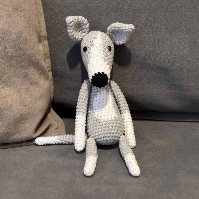 May include: A crocheted dog toy in grey and white. The dog has a long snout, floppy ears, and a black nose. The toy is sitting on a grey surface.