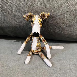 May include: A crocheted dog toy with a long snout and floppy ears. The dog's body is white, brown, and yellow, with white legs and a black nose. The toy is on a grey surface.