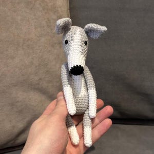 May include: A crocheted greyhound dog toy in grey and white yarn. The dog has a long snout, black nose, and white paws. The handmade doll is a charming, whimsical toy, perfect for children or as a decorative item.