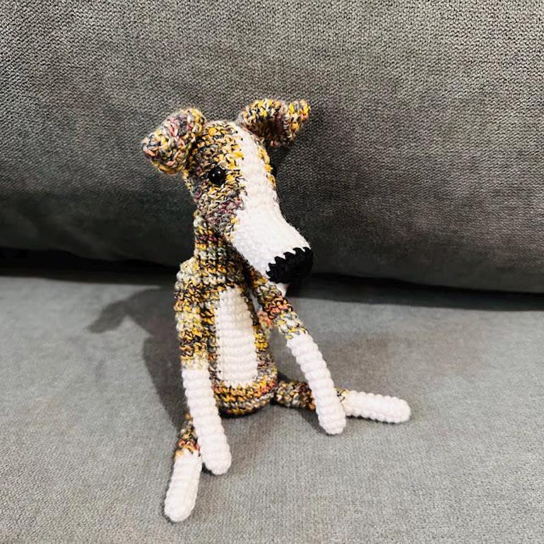 May include: A crocheted dog toy with a long snout and floppy ears. The dog is made from a mix of yellow, brown, and white yarn, with white detailing on the chest, legs, and snout. The nose is black. The dog is sitting on a grey surface.