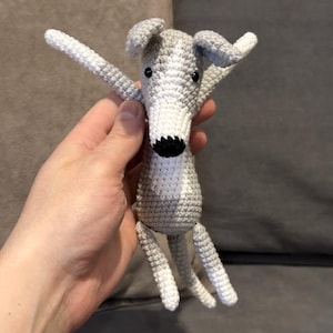 May include: A crocheted greyhound dog toy in grey and white. The dog has a black nose and eyes. The arms and legs are extended. The toy is made of yarn. The dog's body is grey, with a white chest and muzzle.