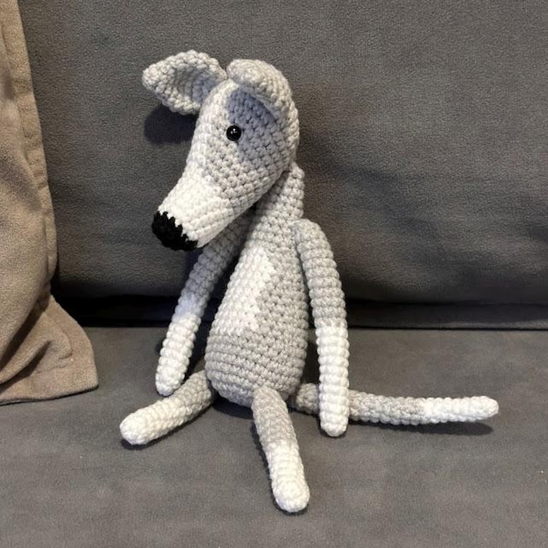 May include: A crocheted dog toy in grey and white. The dog has a long snout, floppy ears, and a small black nose. The toy is sitting on a grey surface.