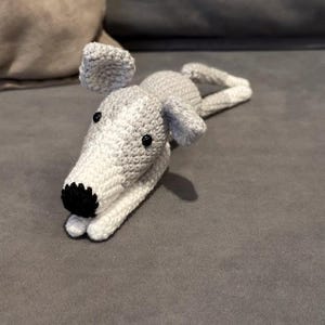 Könnte beinhalten: Ein gehäkeltes Windhund-Spielzeug in liegender Position. Der Hund hat ein weißes Gesicht und eine weiße Brust mit schwarzen Augen und Nase. Die Ohren und der Körper sind grau. Das Spielzeug ist handgefertigt und wirkt weich.