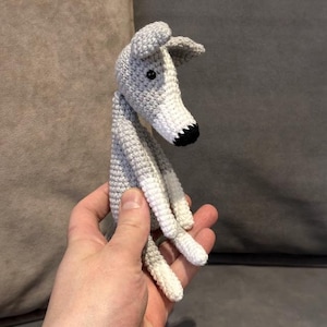 May include: A crocheted greyhound dog toy in grey and white. The dog has a long, thin body and a white snout with a black nose. The ears are grey and the eyes are black. The toy is handmade.