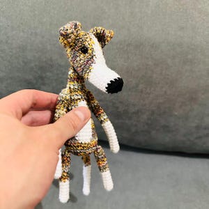 May include: A crocheted dog toy with a long neck and legs. The dog has a white snout and chest, with a black nose. The body is a mix of yellow, brown, and white yarn. The ears and legs are white.