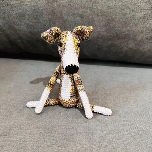 May include: A crocheted dog toy with a long snout, floppy ears, and a black nose. The dog's body is a mix of white, yellow, and brown yarn, with white legs and a white chest. The dog is sitting on a grey surface.