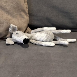 May include: A crocheted dog toy in grey and white, lying on a grey surface. The dog has a black nose and eyes, with floppy ears and a long body. The toy is soft and appears to be made of yarn.
