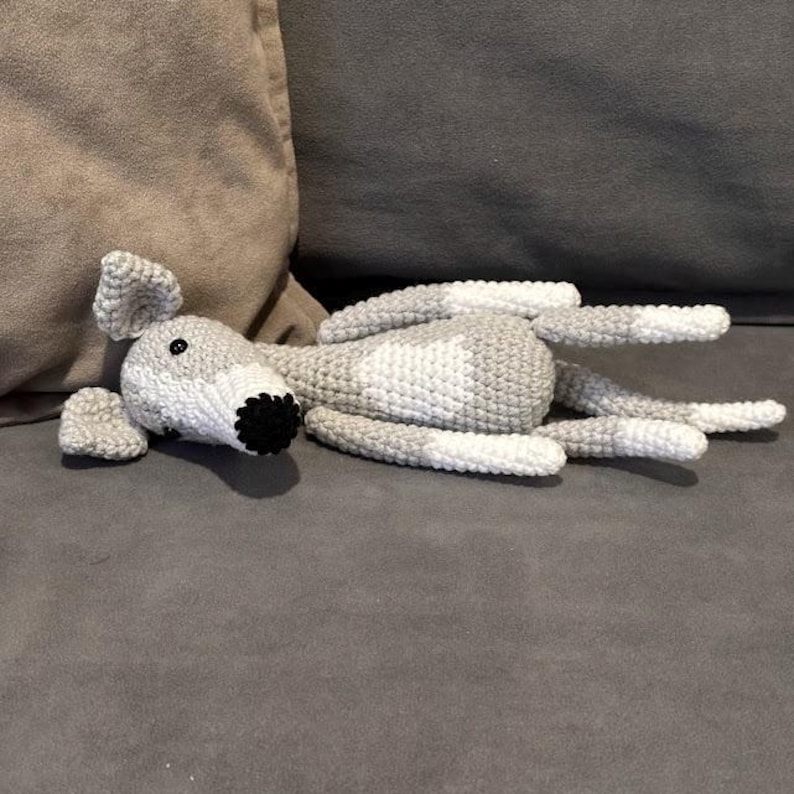 May include: A crocheted dog toy in grey and white, lying on a grey surface. The dog has a black nose and eyes, with floppy ears and a long body. The toy is soft and appears to be made of yarn.