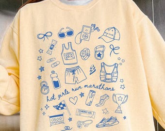 May include: Pale yellow sweatshirt featuring blue marathon-themed illustrations and the text "hot girls run marathons". The design includes running shoes, shorts, and a trophy. A relaxed, casual garment.
