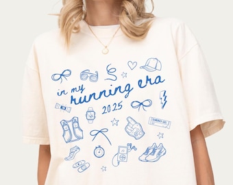 May include: Ivory t-shirt with blue text that says "in my running era 2025" and blue running-themed illustrations.