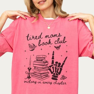 Może przedstawiać: Różowa koszulka z czarnym nadrukiem graficznym, na którym widnieje napis "Tired Moms Book Club" wraz z szkieletową dłonią pokazującą znak rock and roll oraz stosem książek. Tekst pod grafiką brzmi "Victory in every chapter."