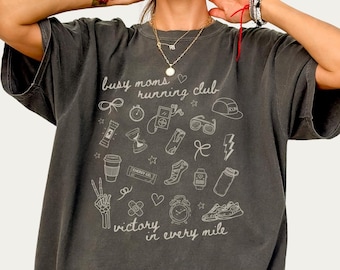 Könnte beinhalten: Ein dunkelgraues Oversize-T-Shirt mit einem weißen Grafikdesign. Die Grafik zeigt den Text "Busy Moms Running Club" und eine Vielzahl von Lauf-Icons, darunter eine Kaffeetasse, eine Stoppuhr, ein Herz und ein Paar Laufschuhe. Der Text "Victory in every mile" ist ebenfalls in der Grafik enthalten.