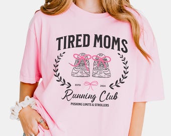 Könnte beinhalten: Hellrosa T-Shirt mit schwarzem Aufdruck "TIRED MOMS Running Club", verziert mit einem Lorbeerkranz und einer Laufschuh-Grafik. Das Shirt trägt auch den Text "PUSHING LIMITS & STROLLERS".