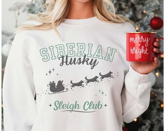 Puede incluir: Sudadera blanca con un gráfico que presenta las palabras "Siberian Husky Sleigh Club" en verde y gris. El diseño incluye un trineo tirado por tres huskys. Se sostiene una taza roja con "merry & bright".