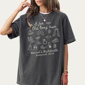 May include: Pepper-colored t-shirt with white graphic text and illustrations. The text reads "in it for the long run" with illustrations of wedding-related items. Below the text is "Hannah's Bachelorette Savannah 2025".