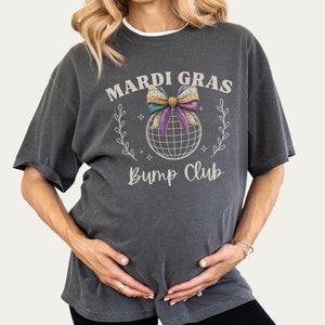 May include: Dark gray t-shirt with the text "MARDI GRAS BUMP CLUB" in a vintage style. The shirt features a graphic of a disco ball with a bow and floral accents. The shirt is a relaxed fit.