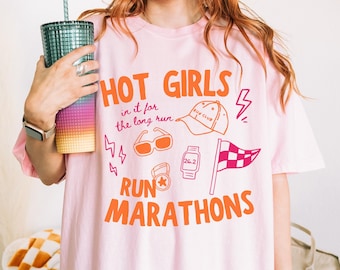 May include: A light pink t-shirt featuring orange and pink graphic text that reads HOT GIRLS RUN MARATHONS and in it for the long run. The design includes illustrations of a baseball cap, sunglasses, a smartwatch, a medal, and a checkered flag.