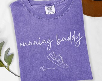 May include: A violet t-shirt featuring the words "running buddy" in a cursive script. Beneath the text is a line drawing of a running shoe and a small heart. The shirt is folded and rests on a white surface.