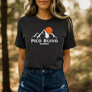 Puede incluir: Camiseta gris oscuro con un diseño de cordillera blanca, un sol naranja y el texto "PICO RUIVO MADEIRA". La camiseta está hecha de un material suave y tiene cuello redondo.