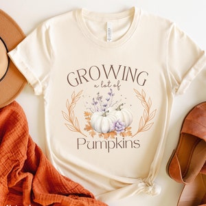 May include: A beige t-shirt with the text "Growing a lot of Pumpkins" in brown lettering. The text is surrounded by a wreath of leaves and flowers with two white pumpkins in the center.