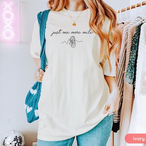 May include: Ivory-colored t-shirt with the text "just one more mile" and a line drawing of a running shoe. The shirt is a relaxed fit and is paired with blue jeans. A blue knitted bag with white hearts is visible.