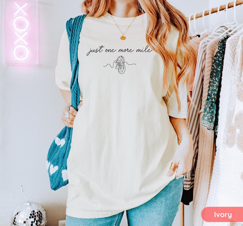 May include: Ivory-colored t-shirt with the text "just one more mile" and a line drawing of a running shoe. The shirt is a relaxed fit and is paired with blue jeans. A blue knitted bag with white hearts is visible.