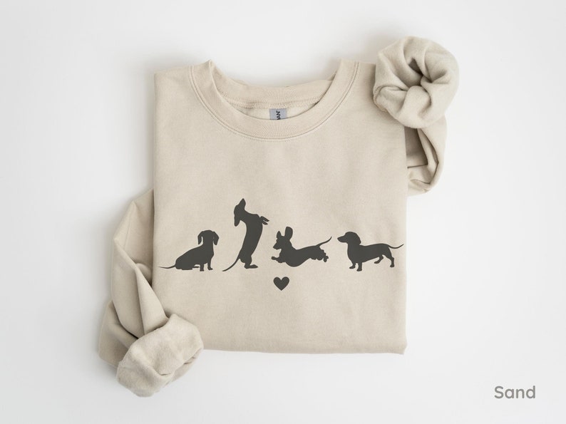 May include: A sand-colored sweatshirt with a graphic of five dachshund silhouettes in various poses. The silhouettes are in black and the sweatshirt has a crew neck.