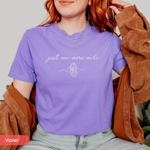 May include: A violet t-shirt with the text "just one more mile" in a cursive font, along with a line drawing of a running shoe. The shirt is short-sleeved and has a crew neck.