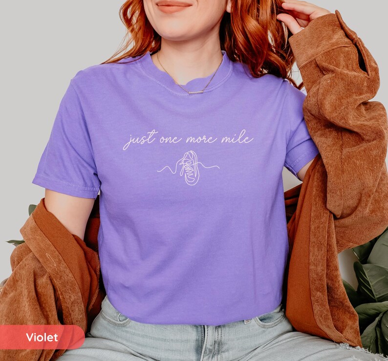 May include: A violet t-shirt with the text "just one more mile" in a cursive font, along with a line drawing of a running shoe. The shirt is short-sleeved and has a crew neck.