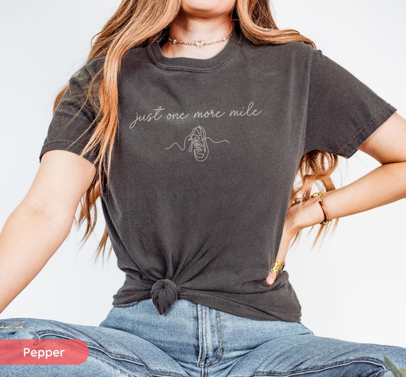 May include: Dark gray t-shirt with the text "just one more mile" and a line drawing of a running shoe. The shirt is knotted at the waist and paired with blue jeans. The shirt is a comfortable, casual style.