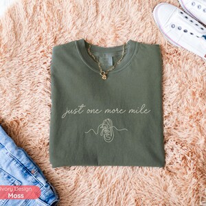 May include: An olive green crew neck t-shirt with the text "just one more mile" and a line drawing of a running shoe. A gold necklace is draped on the shirt. The shirt is on a peach-colored rug.