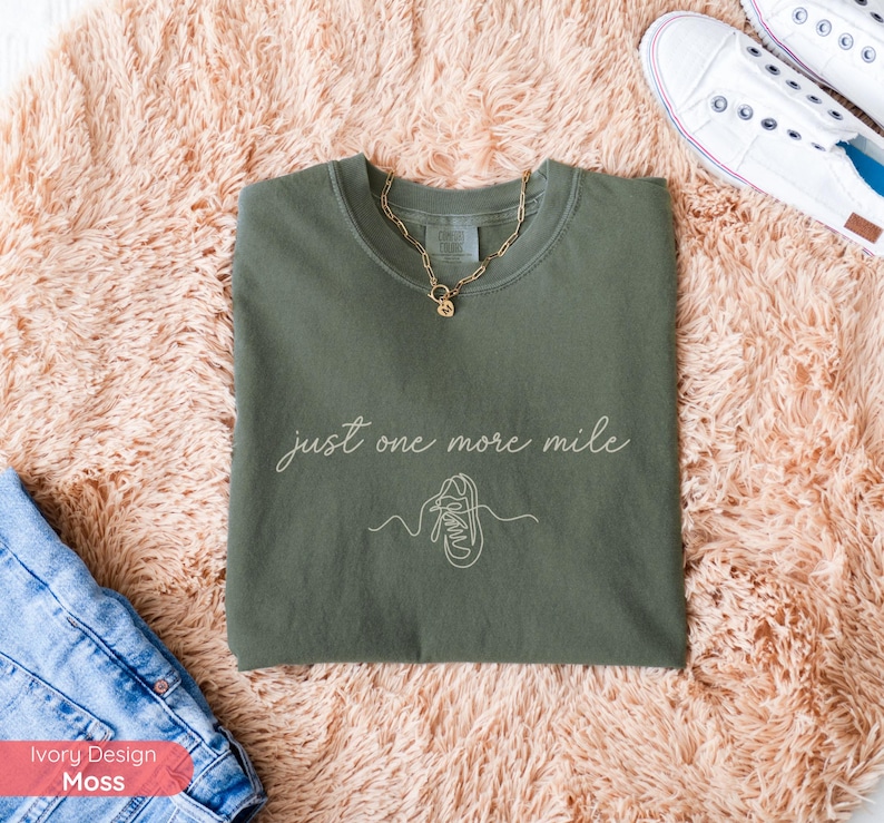 May include: An olive green crew neck t-shirt with the text "just one more mile" and a line drawing of a running shoe. A gold necklace is draped on the shirt. The shirt is on a peach-colored rug.