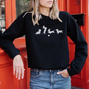 May include: Black sweatshirt with a graphic of five dachshunds in a line. The dachshunds are all facing the same direction and are in different poses. The graphic is in white and is centered on the sweatshirt.