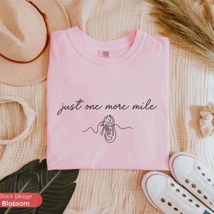 May include: A light pink t-shirt with the text "just one more mile" and a line drawing of a running shoe. The shirt is surrounded by a hat, scrunchie, and white sneakers.