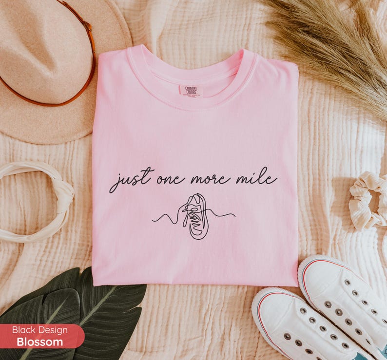 May include: A light pink t-shirt with the text "just one more mile" and a line drawing of a running shoe. The shirt is surrounded by a hat, scrunchie, and white sneakers.