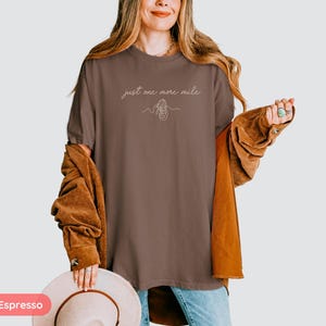 May include: Espresso-colored t-shirt with the text "just one more mile" and a line drawing of a running shoe. The shirt is paired with a brown corduroy jacket and blue jeans. A beige hat is held in the hand.