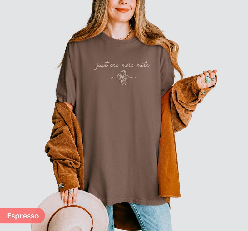 May include: Espresso-colored t-shirt with the text "just one more mile" and a line drawing of a running shoe. The shirt is paired with a brown corduroy jacket and blue jeans. A beige hat is held in the hand.