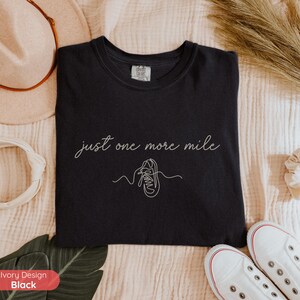 May include: Black t-shirt with the phrase "just one more mile" in cursive script and a line drawing of a running shoe. The shirt is made of a soft material and is a comfortable fit.
