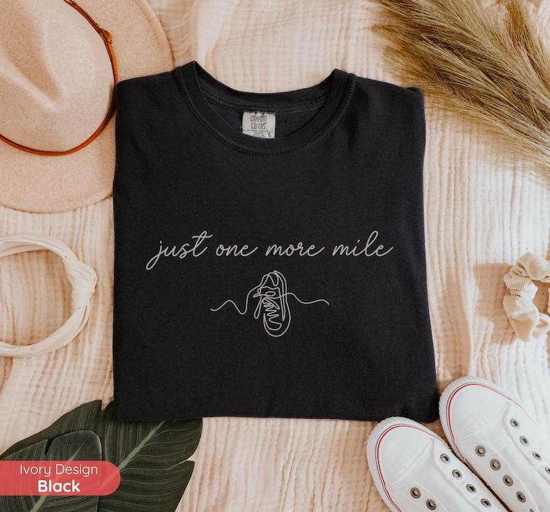 May include: Black t-shirt with the phrase "just one more mile" in cursive script and a line drawing of a running shoe. The shirt is made of a soft material and is a comfortable fit.