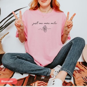 May include: A light pink t-shirt with the text "just one more mile" and a line drawing of a running shoe. The person is wearing blue jeans and white sneakers.