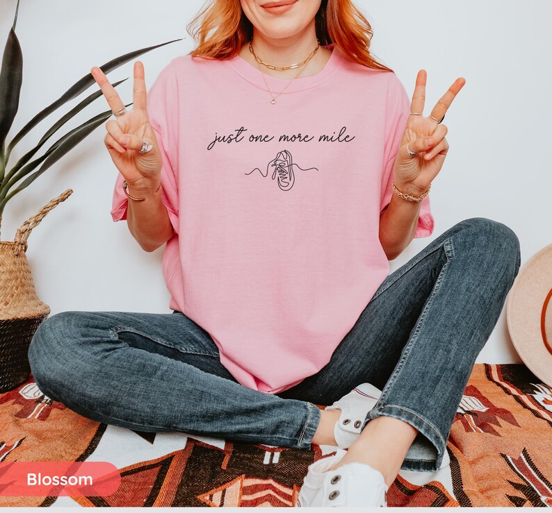 May include: A light pink t-shirt with the text "just one more mile" and a line drawing of a running shoe. The person is wearing blue jeans and white sneakers.