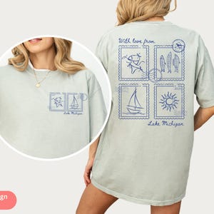 May include: Light green t-shirt with blue stamp-like graphics featuring fish, a sailboat, and a sun. The text "With love from Lake Michigan" is printed on the shirt. The shirt is a relaxed fit.