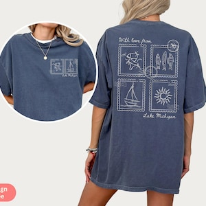 May include: A denim blue tee shirt with a white graphic design. The design includes four postage stamp-like images with drawings of fish, a sailboat, a sun, and a person fishing. The text "With love from Lake Michigan" is also included.