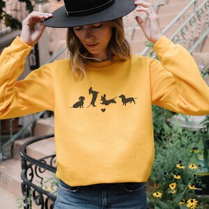 May include: A yellow sweatshirt with a graphic of four dachshund silhouettes. The dachshunds are in various poses, including sitting, standing, and running. The graphic includes a small heart.